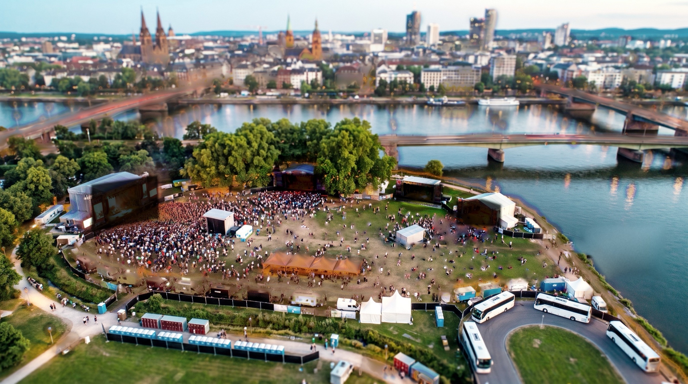 A riverside city park from above in the morning: stages dark, the first people arriving, buses parked by the gate