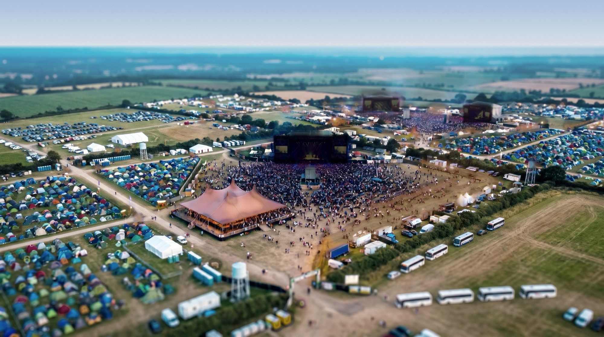 The camping festival from above in cool morning light: tents, the dark Main Stage, coaches by the gate.