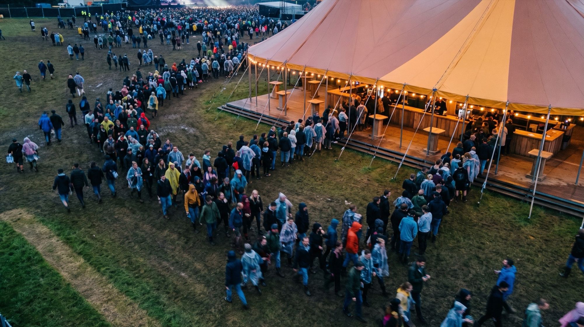 Seen from above, people in ponchos queue in long lanes into a busy copper big-top bar.